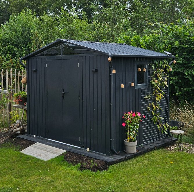 Antraciet metalen tuinhuis met een bloeiende plant en lampjes in de tuin van een klant